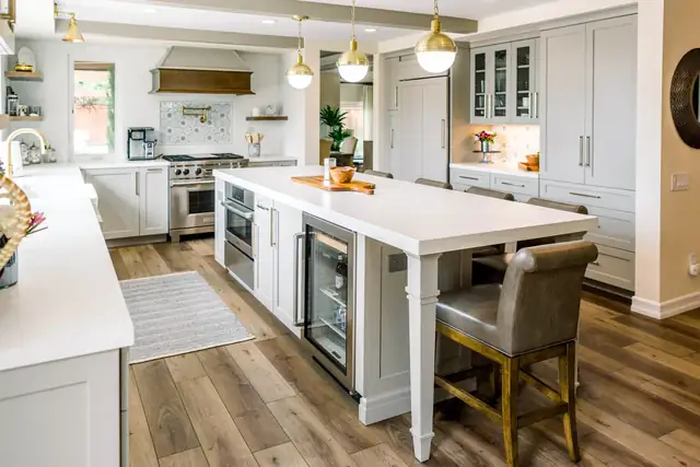 Farmhouse kitchen by Kitchen Barn with white cabinets, a white island with a beverage fridge, brass globe pendants and a wood range hood