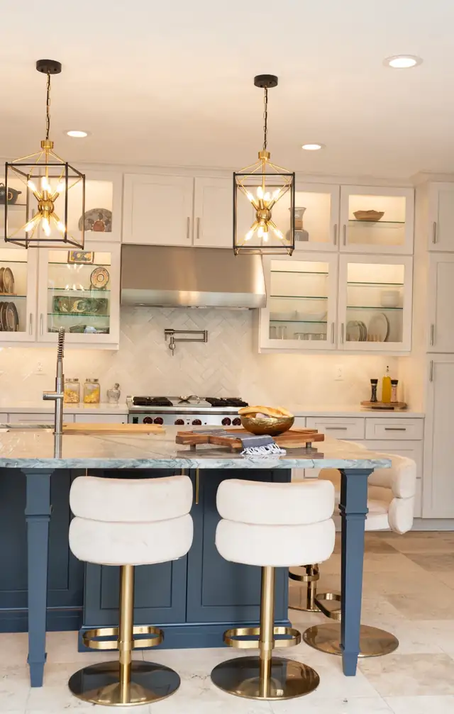 White kitchen with a navy island, cream counter stools, glass-front upper cabinets and brass lantern pendants