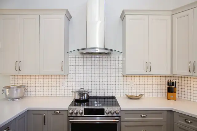 Remodeled kitchen with white upper cabinets, gray lower cabinets, dot-pattern tile backsplash and a stainless chimney hood